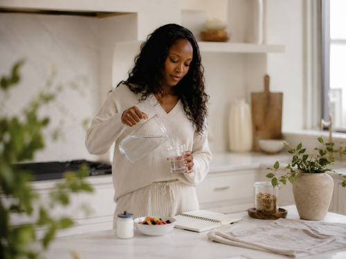 A Black woman pouring water in a calm kitchen beside fruit, an open notebook and soft morning light.