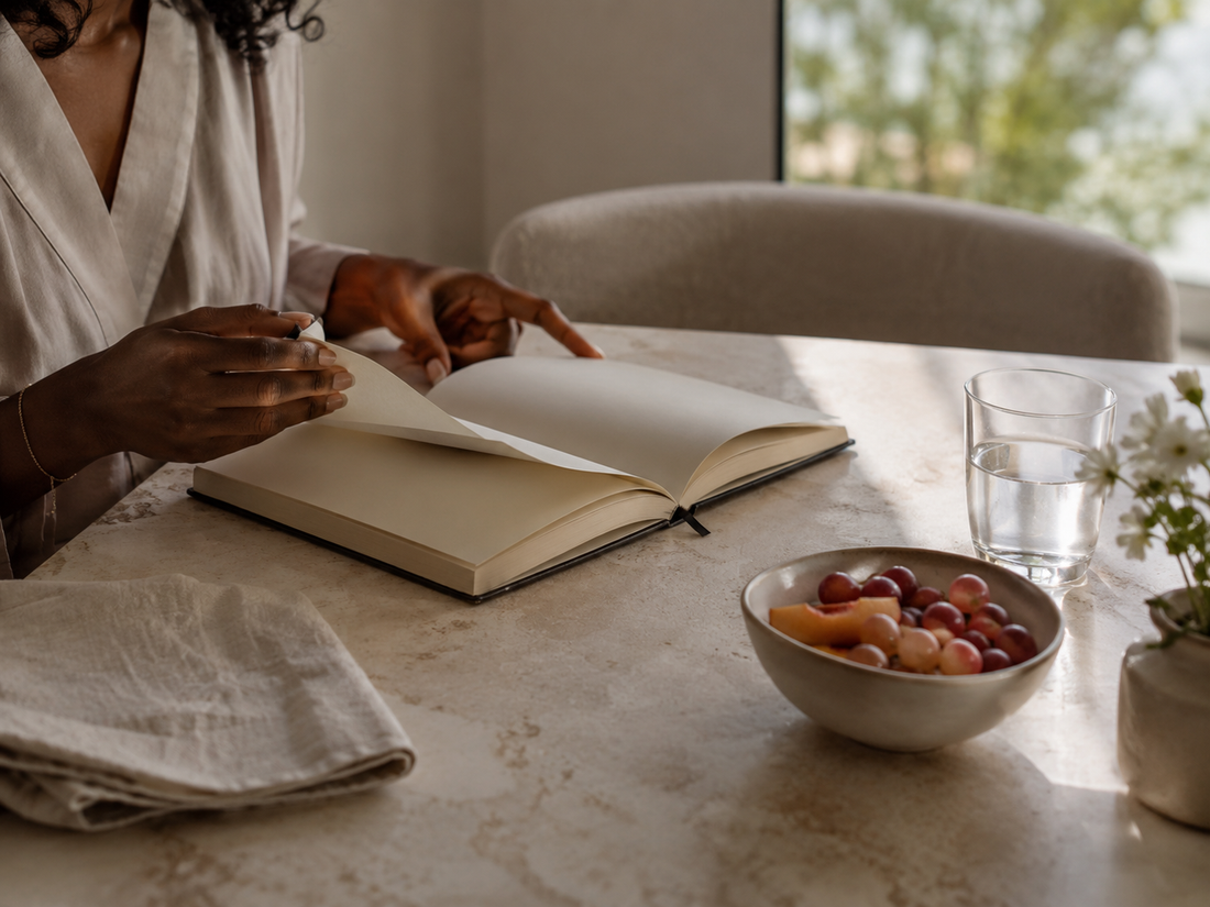 A Black woman gently turning the pages of an open planner beside water, fruit and soft morning light.